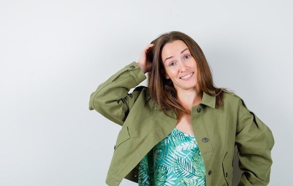 Giovane donna che si gratta la testa mentre sorride in giacca verde e sembra indecisa, vista frontale.