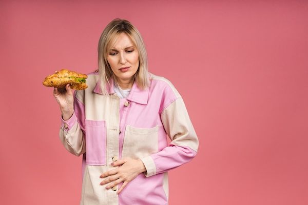 Giovane donna bella con dolore addominale, isolata su sfondo rosa, che tiene in mano del cibo spazzatura. Concetto di intossico.