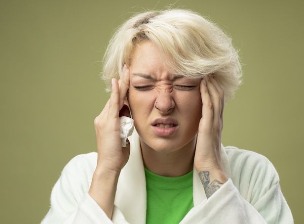 Donna malata con i capelli corti che si sente male, toccandosi le tempie a causa di meningite, in piedi su uno sfondo chiaro.