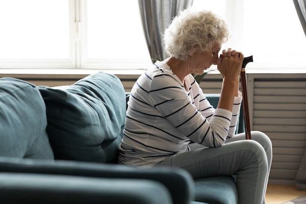 Vista laterale di un'anziana nonna matura e stanca seduta sul divano con un bastone di legno tra le mani, con sintomi iniziali di Alzheimer. Pensionata infelice, anziana che soffre di disabilità, sola nel soggiorno.