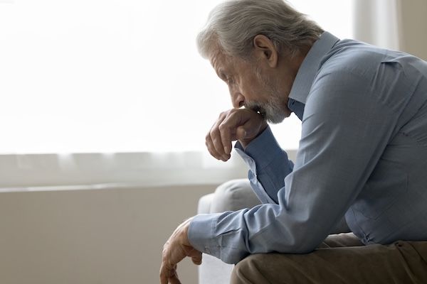 Anziano pensionato depresso seduto sul divano di casa sua, con lo sguardo abbassato, pensando ai problemi legati all'assistenza sanitaria durante la pensione. Depressione, senso di solitudine, ansia, sofferenza per apatia, ansia e solitudine, sintomi di Alzheimer.