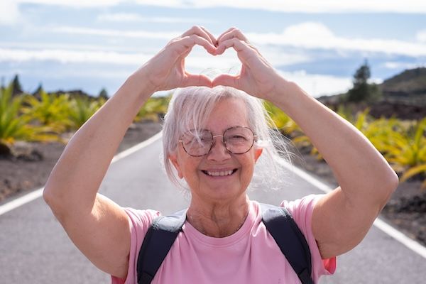 Ritratto di una donna anziana allegra con zaino, mentre cammina all'aperto su una strada deserta, formando un gesto a forma di cuore con le mani per dare un messaggio di prevenzione dell'aritmia cardiaca per gli anziani