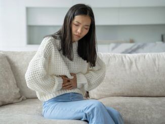 Una donna asiatica con una camicetta bianca è seduta sul divano e sente un forte dolore addominale per via dell'influenza intestinale