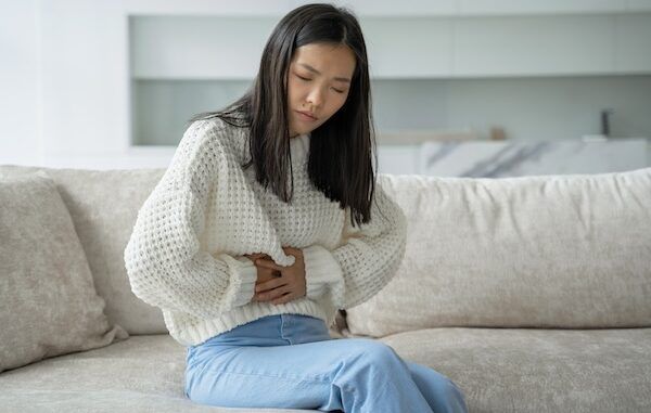 Una donna asiatica con una camicetta bianca è seduta sul divano e sente un forte dolore addominale per via dell'influenza intestinale
