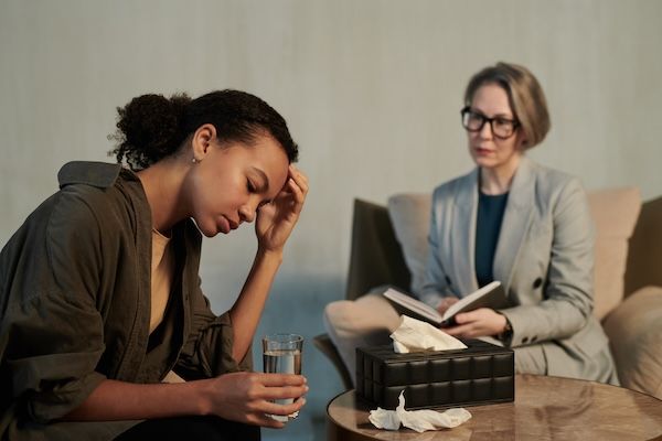 Giovane paziente afroamericana triste con bicchiere d'acqua di fronte allo psicoterapeuta o psicologo