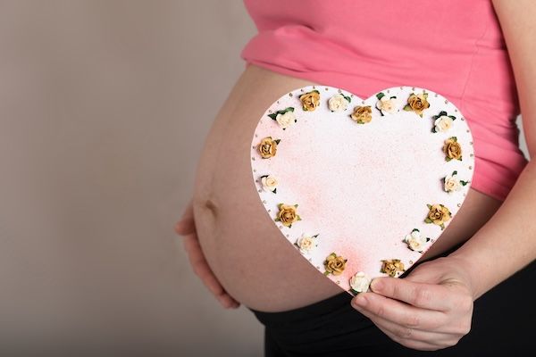 Giovane donna incinta tra i 30 e i 35 anni tiene un cuore di carta decorato. Vista da vicino.