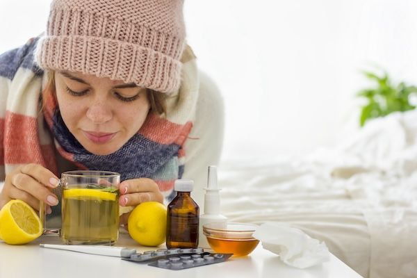 Giovane donna ben vestita sdraiata sul letto, mentre beve tè caldo con limone e prende un altro medicinale per abbassare la febbre alta