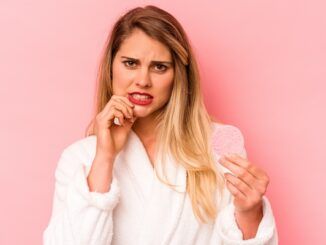 Giovane donna caucasica che tiene una spugna per il viso, isolata su sfondo rosa, si morde nervosamente le unghie e appare molto ansiosa a causa dell'ascesso gengivale scoppia da solo