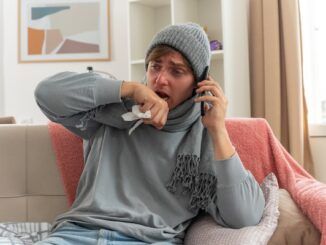 Giovane malato di bronchite senza febbre con una sciarpa intorno al collo e un cappello invernale, che tiene una mano vicino alla bocca e parla al telefono seduto sul divano in soggiorno.