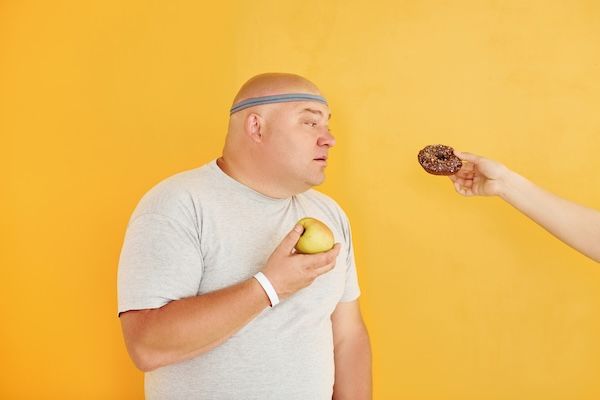 Mela o ciambella? Uomo simpatico e sovrappeso con una cravatta sportiva in testa su sfondo giallo sui cibi da evitare per chi ha il colesterolo alto