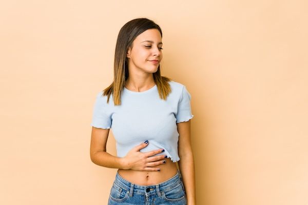 Giovane donna isolata su sfondo beige tocca la pancia, motilità intestinale, sorride dolcemente, concetto di cibo e soddisfazione.