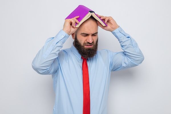 Uomo barbuto con cravatta rossa e camicia blu che tiene un libro sopra la testa con un aspetto stanco e irritato perché non sa cos'è l'adhd