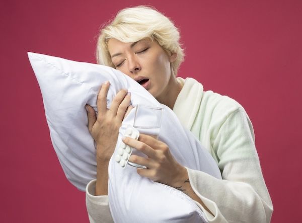 Donna malata con capelli corti che abbraccia un cuscino, soffre di apnea notturna e si sente male, tiene un bicchiere d'acqua e medicine, in piedi davanti a un muro rosa