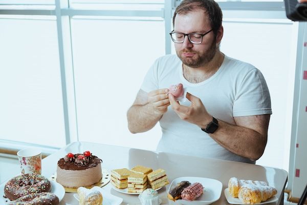 Un uomo in sovrappeso che mangia un dessert davanti a un tavolo pieno di cibo dolce invece di fare una dieta per abbassare il colesterolo alto
