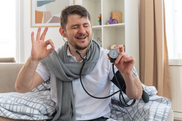 Uomo malato soddisfatto con una sciarpa intorno al collo che misura la pressione con uno sfigmomanometro e fa il gesto di "ok", seduto sul divano nel soggiorno per una prevenzione cardiovascolare ottimale