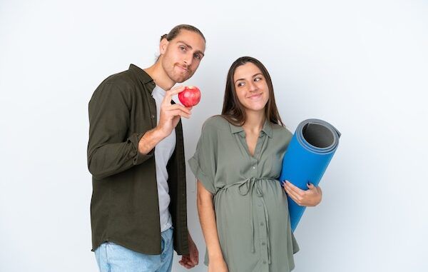 la prevenzione cardiovascolare inizia da giovani. Giovane coppia caucasica isolata su sfondo bianco, con la donna incinta che tiene in mano una mela e partecipa a lezioni di yoga.