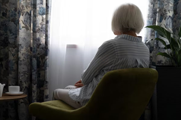 Una donna anziana, seduta con le spalle rivolte alla telecamera, guarda fuori da una finestra coperta da una